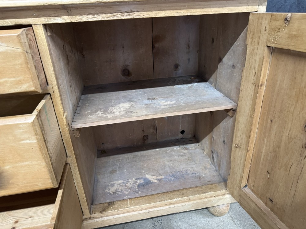 A small antique pine dresser base featuring three drawers on the left, a single panelled cupboard on the right, wooden knobs, turned bun feet, and a shaped back gallery shelf. View V