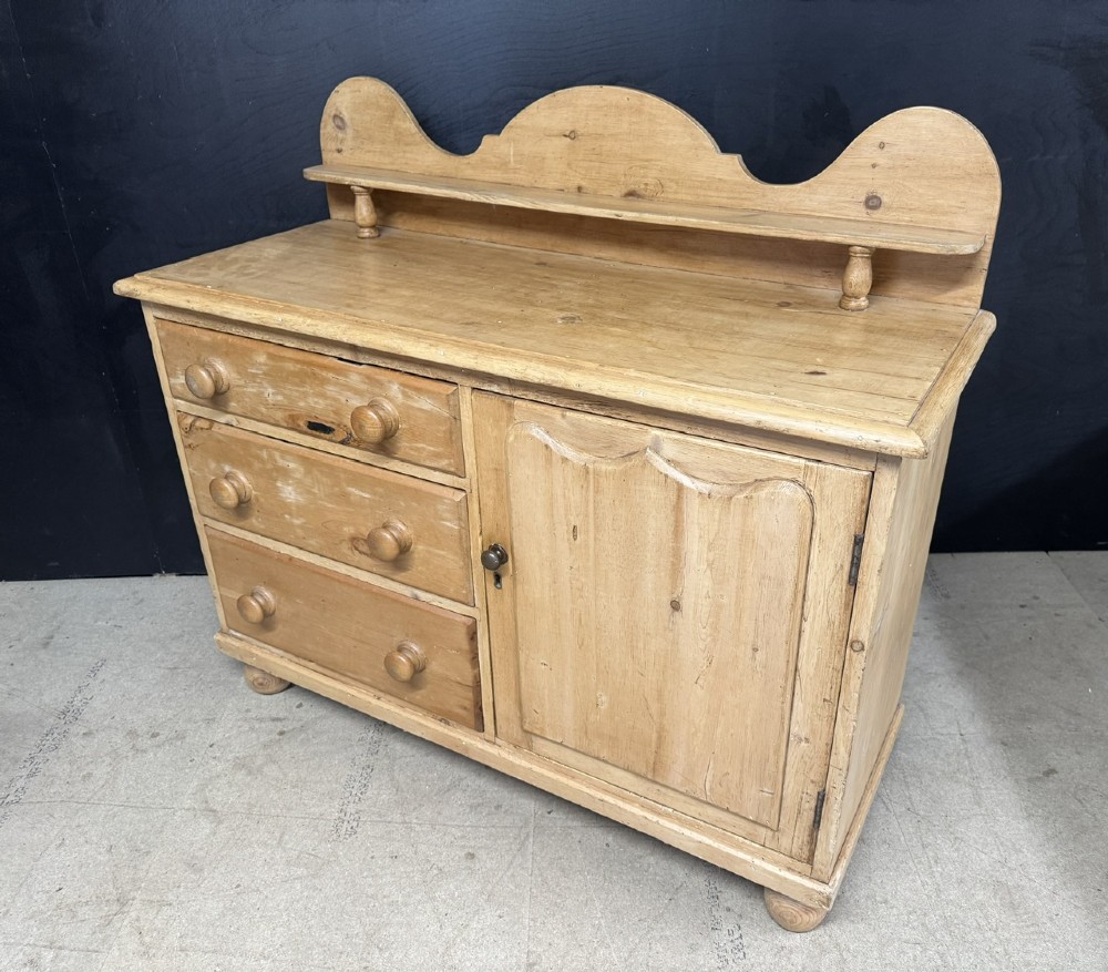 A small antique pine dresser base featuring three drawers on the left, a single panelled cupboard on the right, wooden knobs, turned bun feet, and a shaped back gallery shelf. View VI