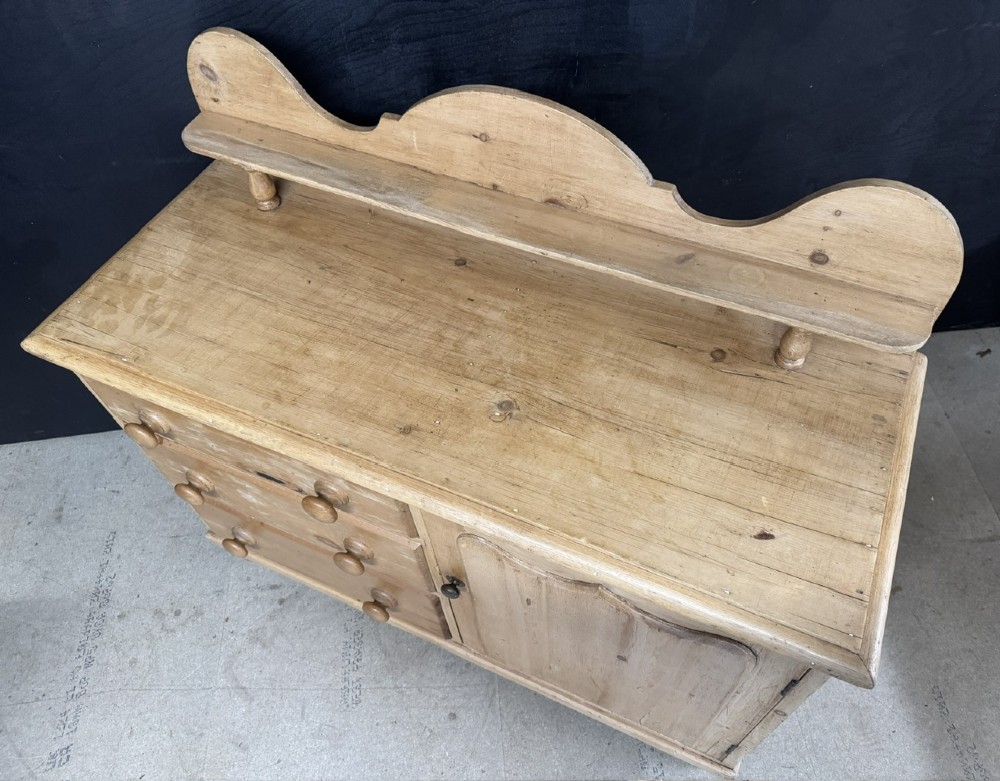 A small antique pine dresser base featuring three drawers on the left, a single panelled cupboard on the right, wooden knobs, turned bun feet, and a shaped back gallery shelf. View VII