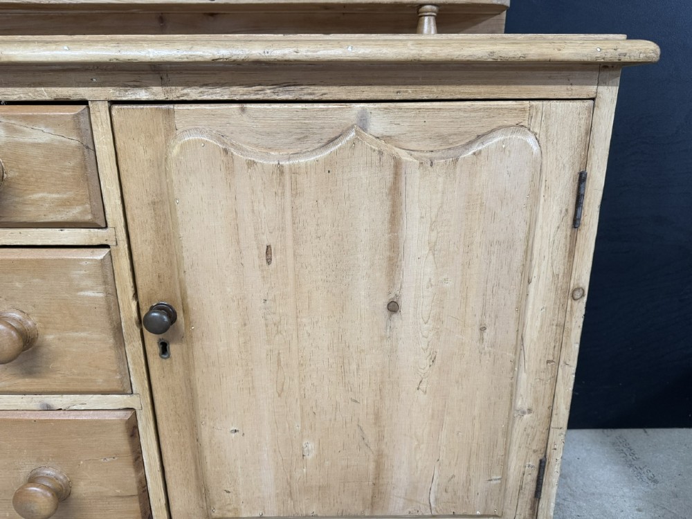 A small antique pine dresser base featuring three drawers on the left, a single panelled cupboard on the right, wooden knobs, turned bun feet, and a shaped back gallery shelf. View X