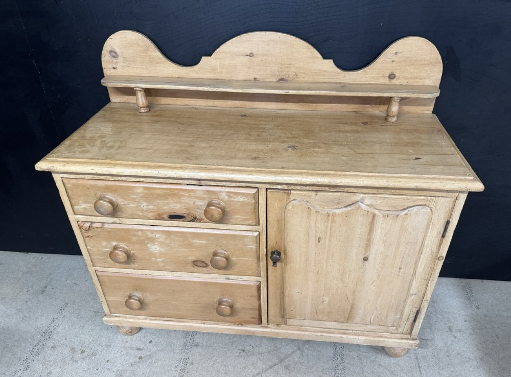 A small antique pine dresser base featuring three drawers on the left, a single panelled cupboard on the right, wooden knobs, turned bun feet, and a shaped back gallery shelf. View XI