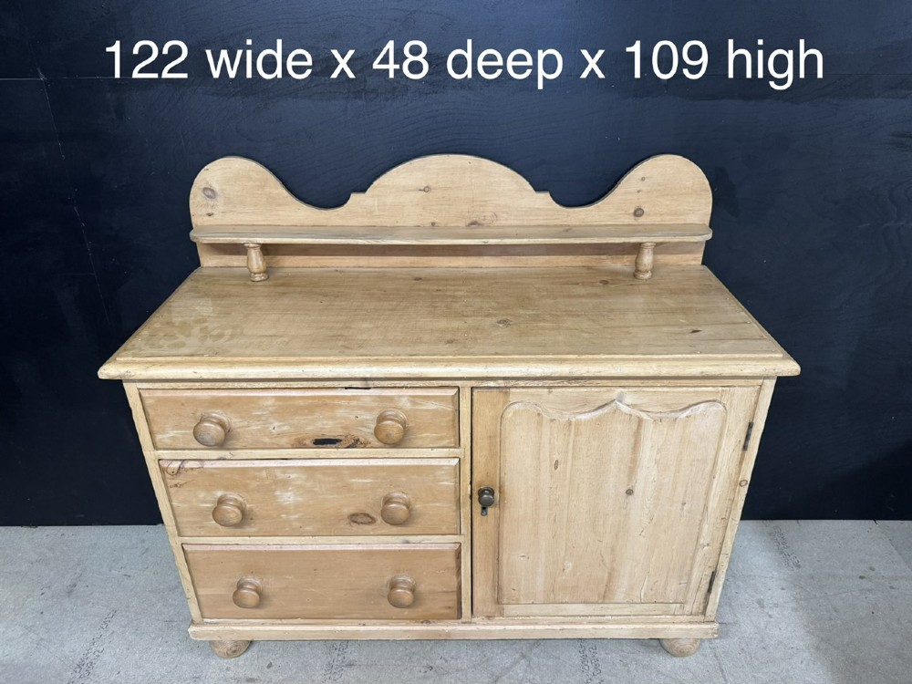 A small antique pine dresser base featuring three drawers on the left, a single panelled cupboard on the right, wooden knobs, turned bun feet, and a shaped back gallery shelf. View XIII