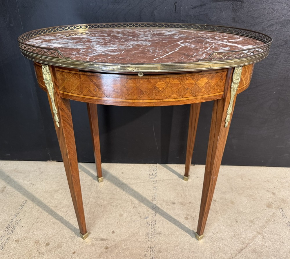 An oval French side table featuring a red veined marble top with a pierced brass gallery, geometric parquetry apron, and four tapered legs adorned with brass mounts.