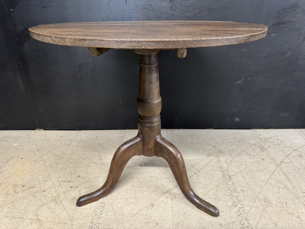 A circular Georgian oak tilt-top table positioned upright on a central turned column with three curved tripod legs. View VI