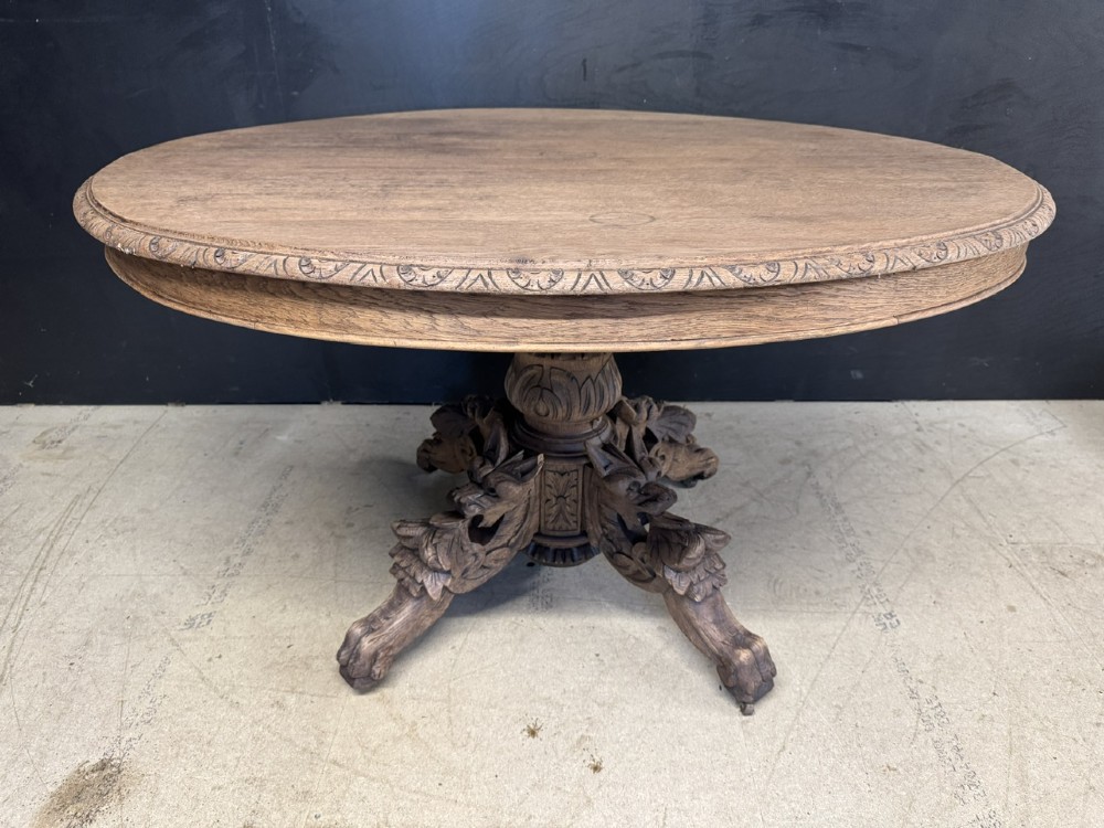A round bleached oak dining table featuring an intricately carved apron and a central pedestal supported by four beast-head scrolled legs.