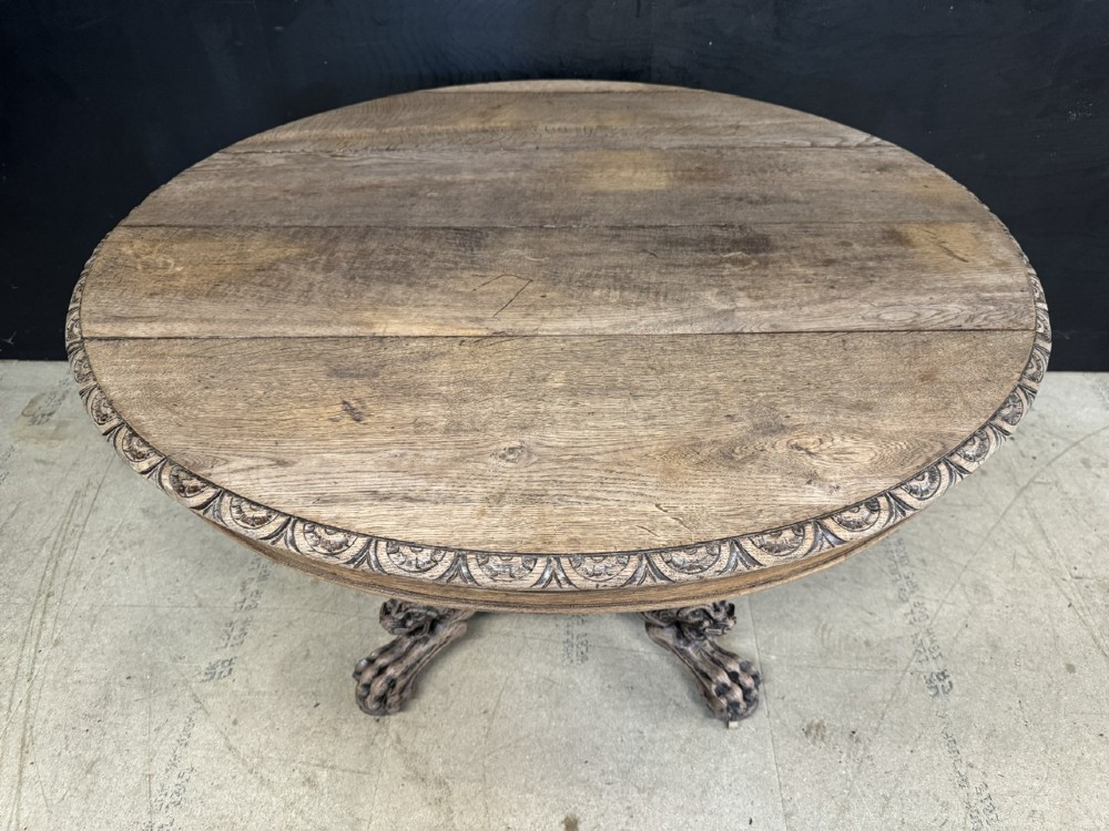 A round bleached oak dining table featuring an intricately carved apron and a central pedestal supported by four beast-head scrolled legs. View III