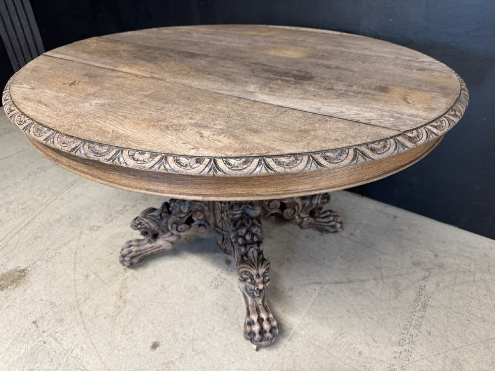 A round bleached oak dining table featuring an intricately carved apron and a central pedestal supported by four beast-head scrolled legs. View X