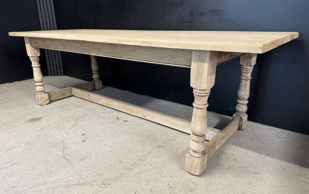 A large light oak farmhouse dining table featuring a solid plank top, turned legs, and an H-shaped stretcher base. View X