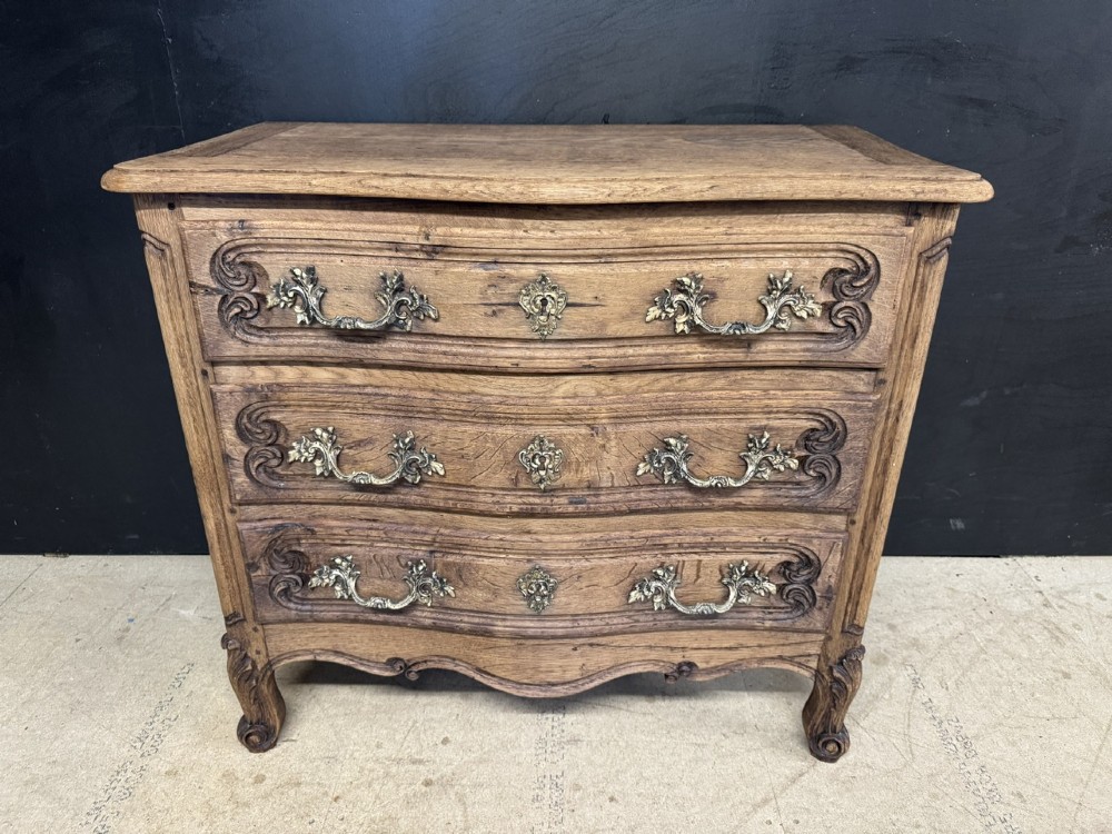 A French provincial bleached oak commode featuring three drawers with carved decorative mouldings and cast metal handles.