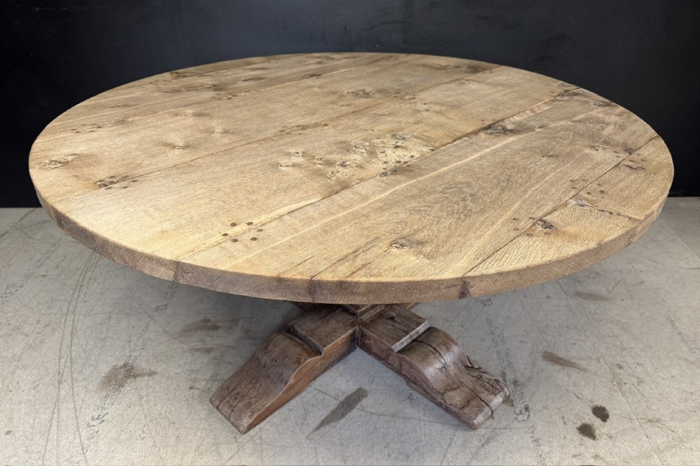 A large round oak dining table featuring a planked top and a substantial wooden cross-pedestal base.