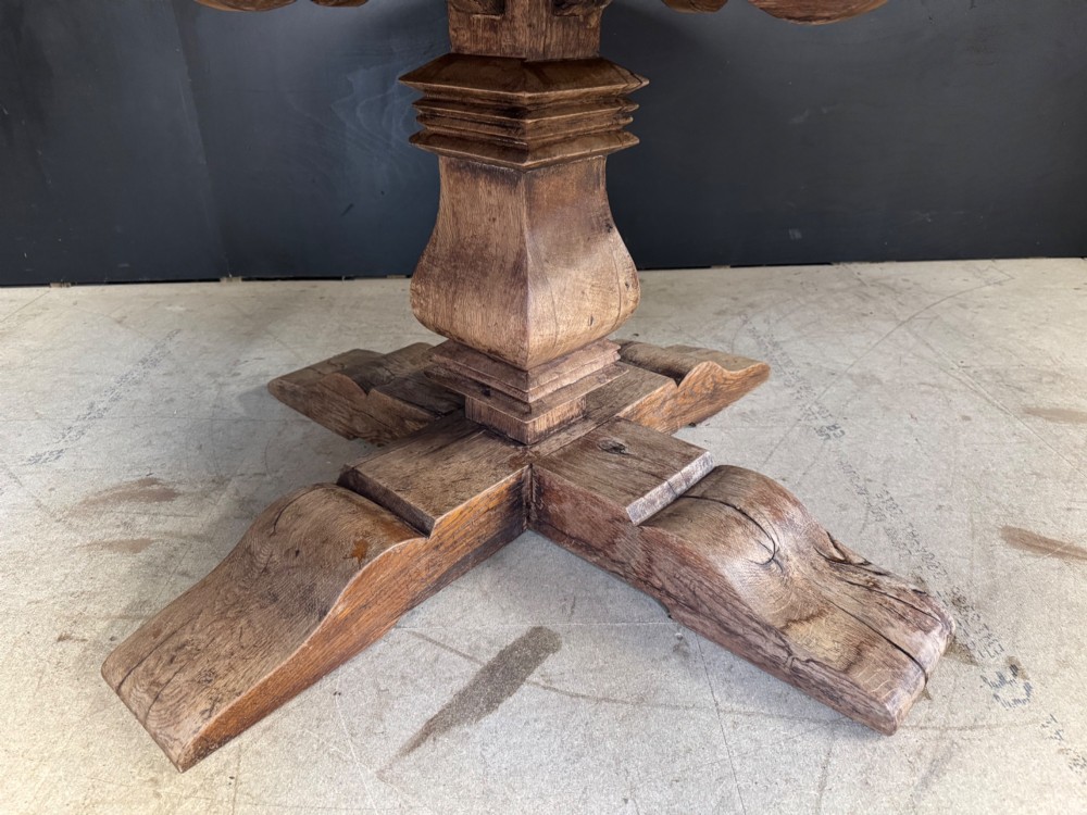 A large round oak dining table featuring a planked top and a substantial wooden cross-pedestal base. View V