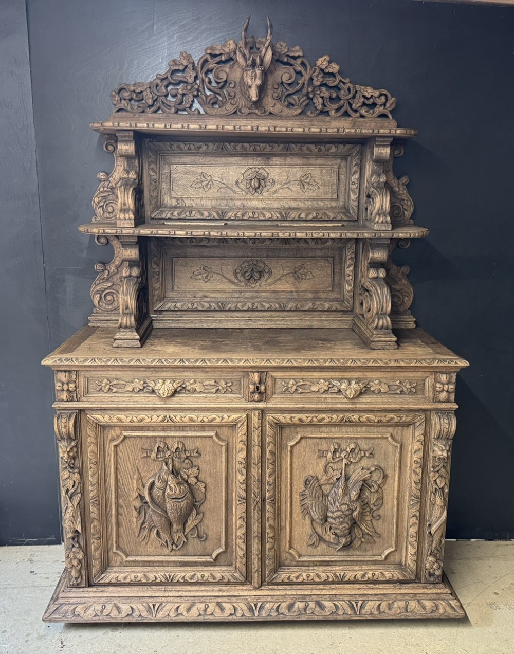 A dark oak French hunting sideboard featuring a carved stag head top pediment, open middle shelves, and lower cabinet doors decorated with carved game birds.