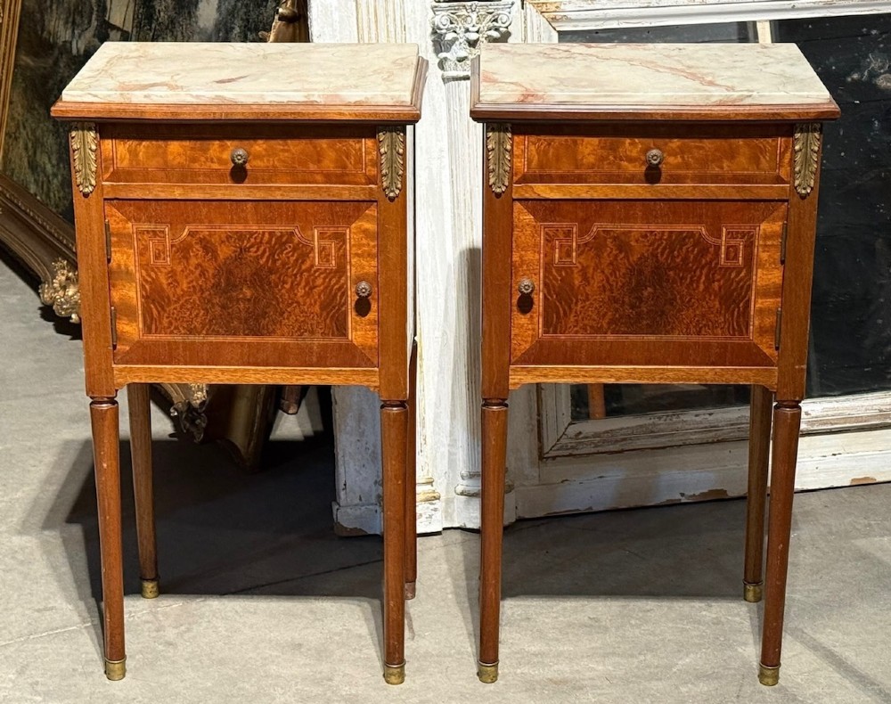 A pair of French Louis XVI style wooden bedside cabinets featuring marble tops, single drawers, inlaid cupboard doors, and gilt brass mounts on slender tapered legs.