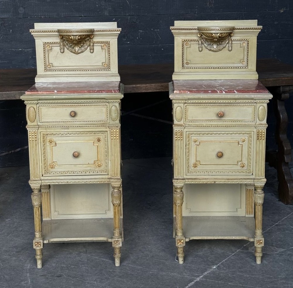 A matching pair of painted French neoclassical bedside cabinets featuring red marble tops, carved bracket superstructures, gold leaf detailing, drawers, and open lower shelves.