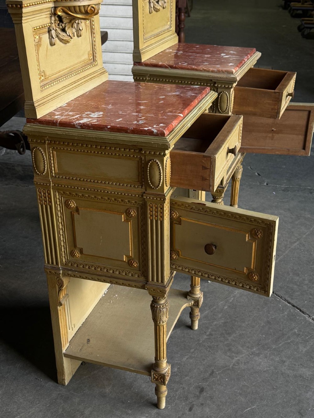 A matching pair of painted French neoclassical bedside cabinets featuring red marble tops, carved bracket superstructures, gold leaf detailing, drawers, and open lower shelves. View VII