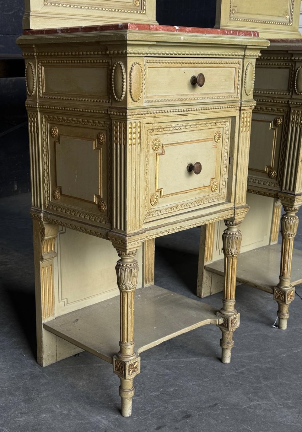 A matching pair of painted French neoclassical bedside cabinets featuring red marble tops, carved bracket superstructures, gold leaf detailing, drawers, and open lower shelves. View VIII