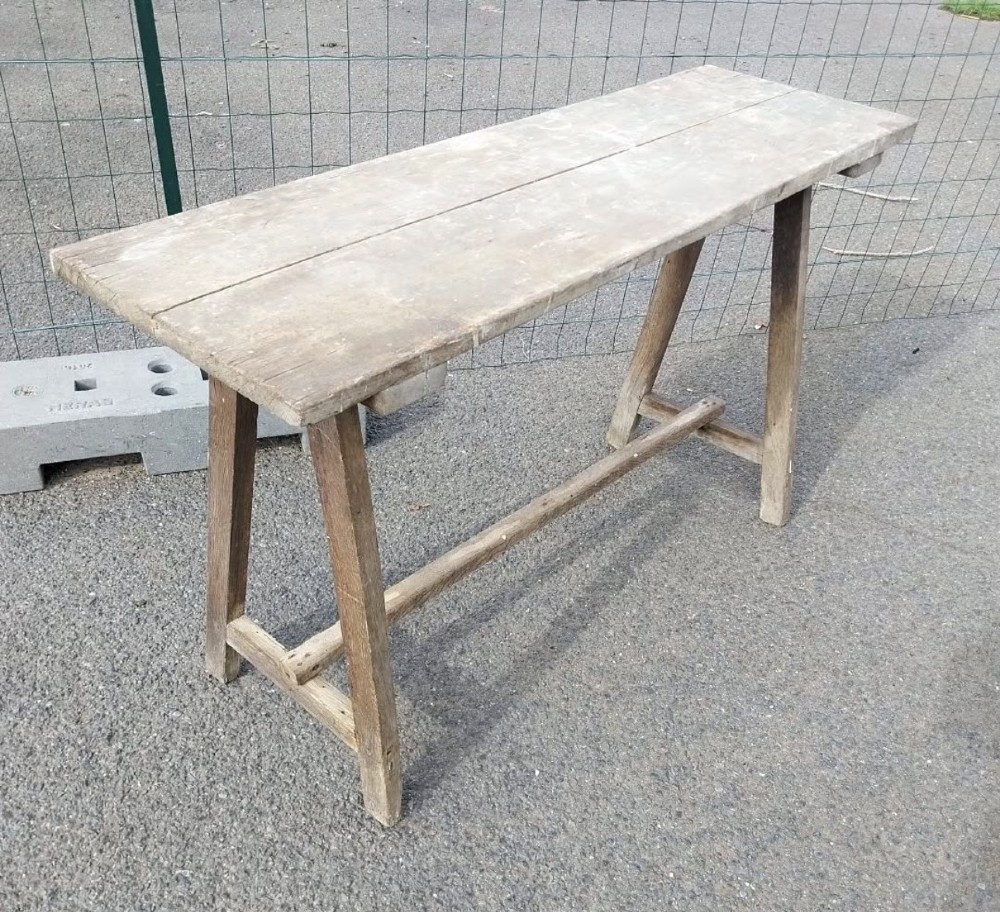 A rustic weathered wooden French laundry table featuring a plank top supported by angled A-frame trestle legs and a central stretcher.