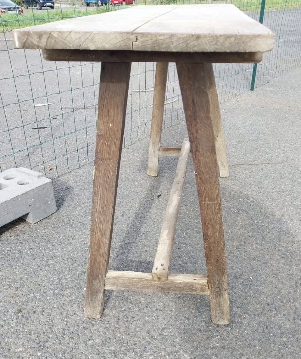A rustic weathered wooden French laundry table featuring a plank top supported by angled A-frame trestle legs and a central stretcher. View V