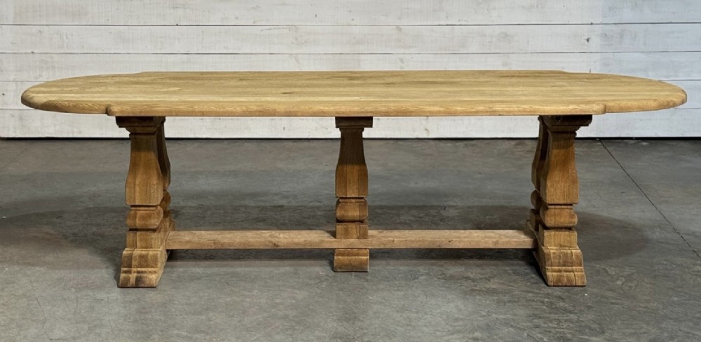 A large rectangular bleached oak dining table featuring carved trestle supports, a central low connecting stretcher, and rounded corner edges.