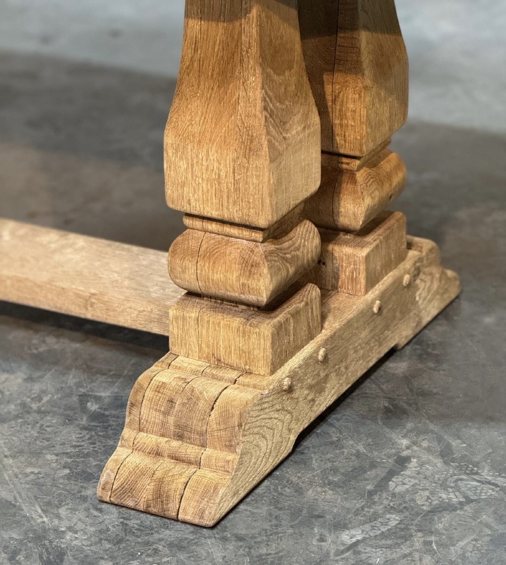 A large rectangular bleached oak dining table featuring carved trestle supports, a central low connecting stretcher, and rounded corner edges. View V