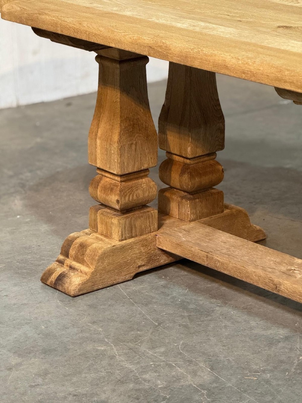 A large rectangular bleached oak dining table featuring carved trestle supports, a central low connecting stretcher, and rounded corner edges. View VI