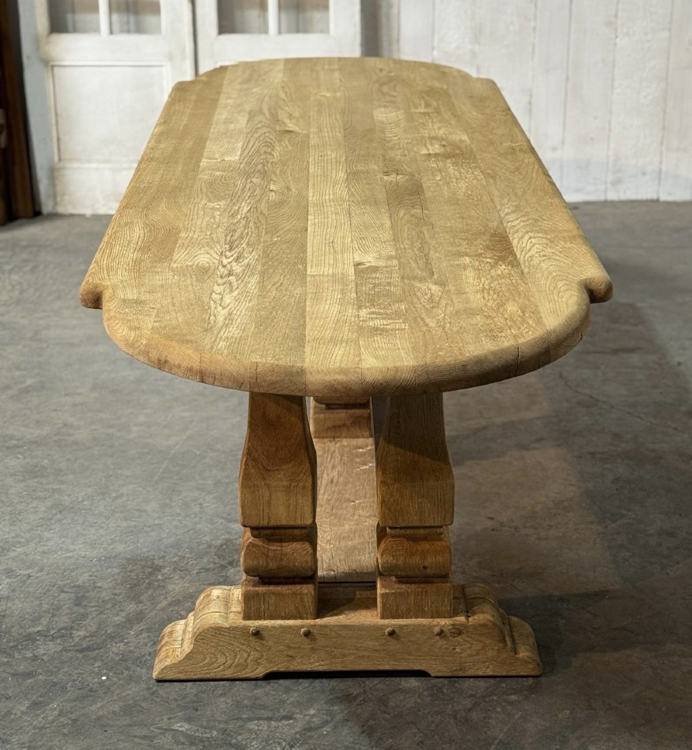 A large rectangular bleached oak dining table featuring carved trestle supports, a central low connecting stretcher, and rounded corner edges. View VIII