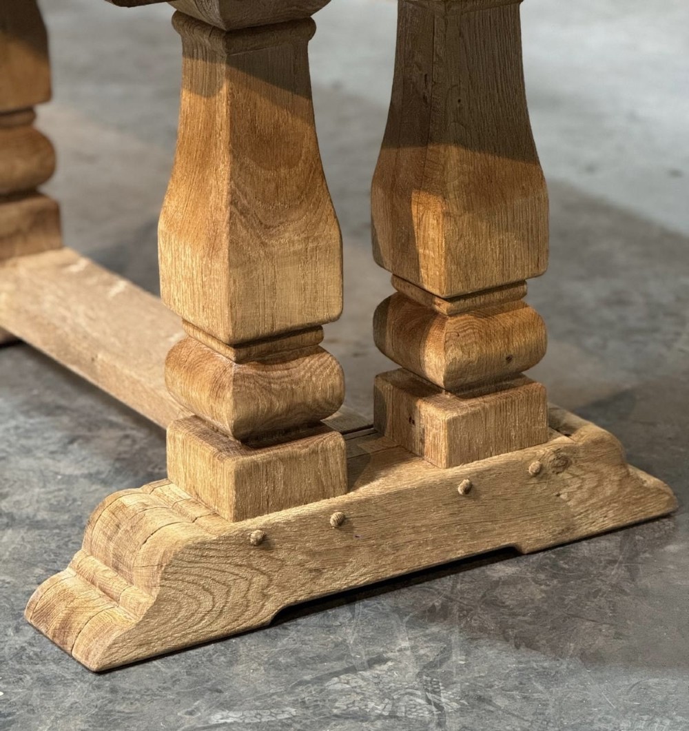 A large rectangular bleached oak dining table featuring carved trestle supports, a central low connecting stretcher, and rounded corner edges. View IX