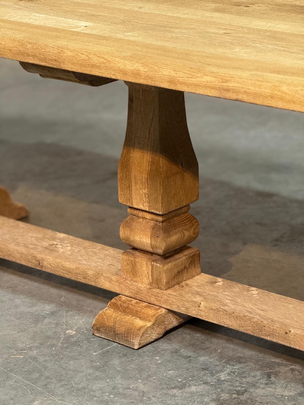 A large rectangular bleached oak dining table featuring carved trestle supports, a central low connecting stretcher, and rounded corner edges. View X