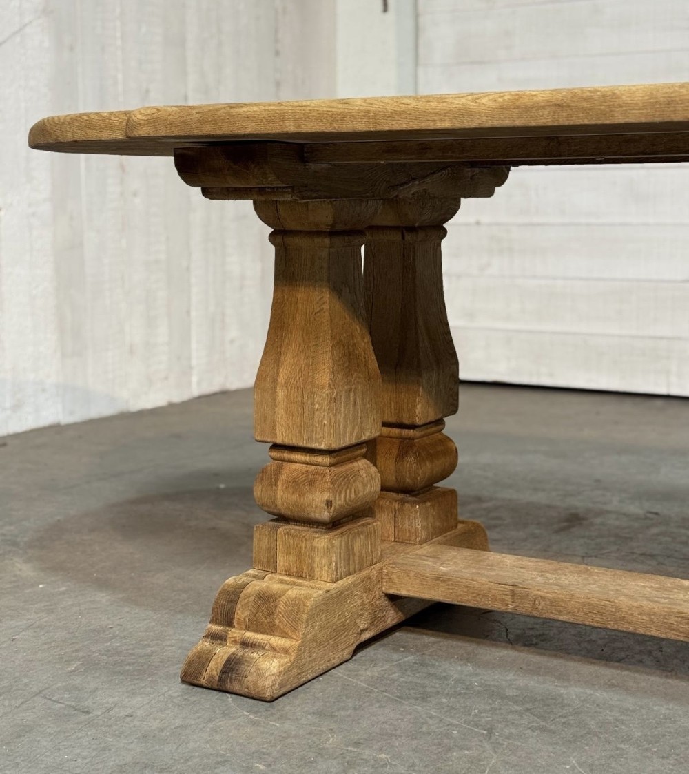 A large rectangular bleached oak dining table featuring carved trestle supports, a central low connecting stretcher, and rounded corner edges. View XI