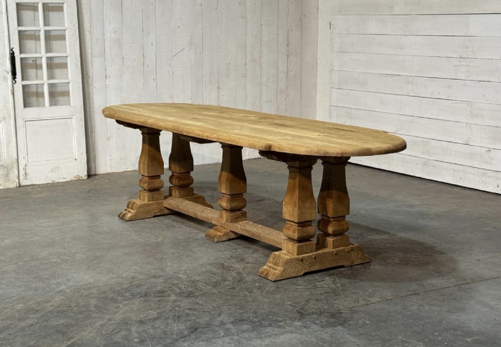 A large rectangular bleached oak dining table featuring carved trestle supports, a central low connecting stretcher, and rounded corner edges. View XII