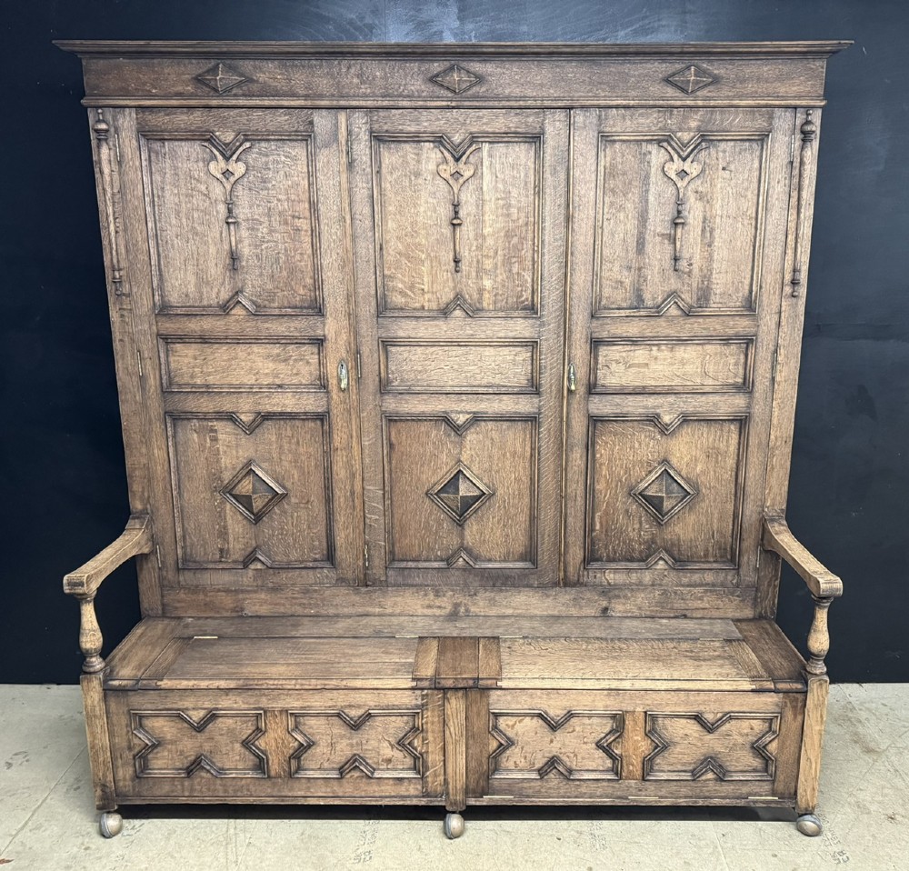 A large carved oak hall bench featuring a high back with three cupboard doors, geometric diamond panelling, and a lift-top storage seat.