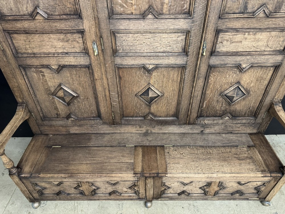 A large carved oak hall bench featuring a high back with three cupboard doors, geometric diamond panelling, and a lift-top storage seat. View V