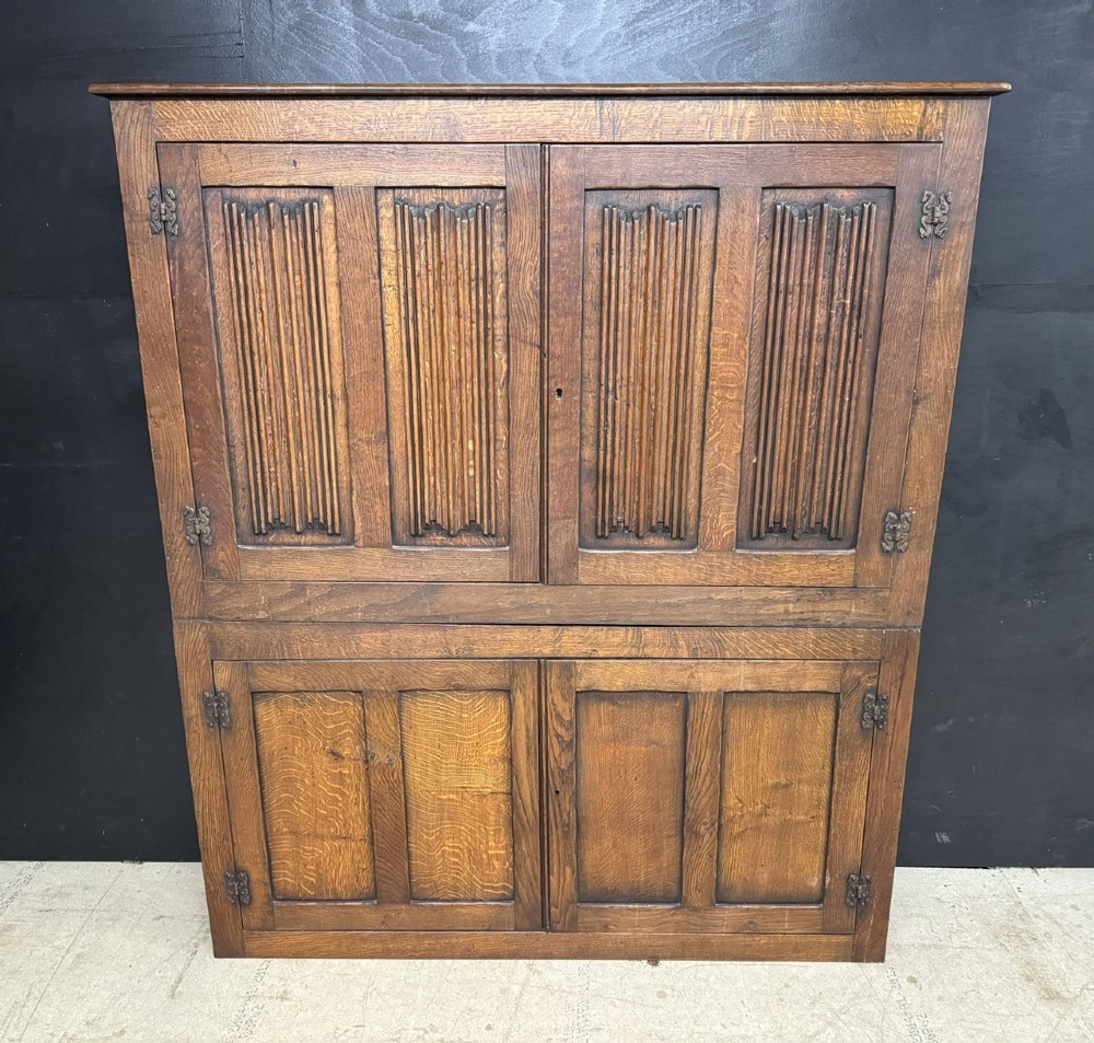 A narrow dark oak hall cupboard featuring upper carved linenfold doors and lower plain panel doors with iron hinges.