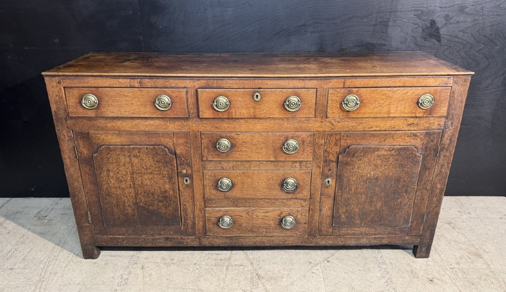 A Georgian Welsh oak dresser base with multiple drawers and two panelled cupboard doors featuring brass drop handles.