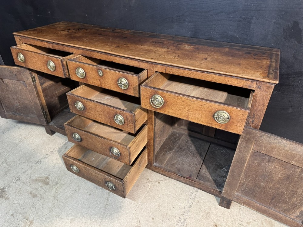A Georgian Welsh oak dresser base with multiple drawers and two panelled cupboard doors featuring brass drop handles. View V