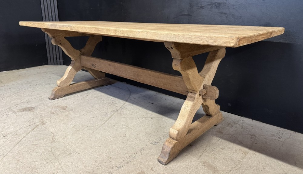 A rustic light oak dining table featuring a thick plank top supported by X-frame trestle legs and a pegged central stretcher.
