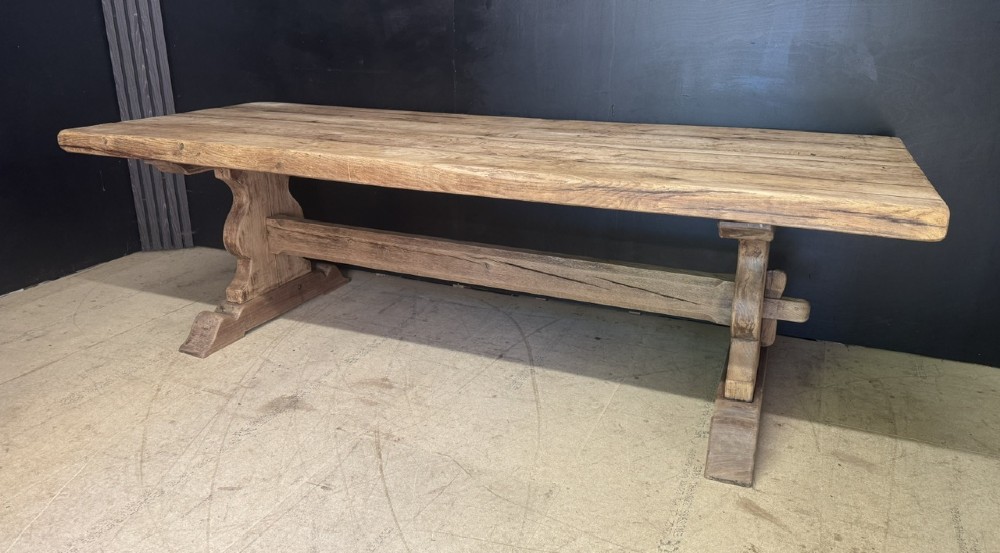 A large oak farmhouse dining table featuring a solid plank top resting on trestle end supports and a keyed stretcher. View II