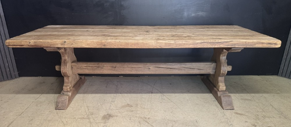 A large oak farmhouse dining table featuring a solid plank top resting on trestle end supports and a keyed stretcher. View VII