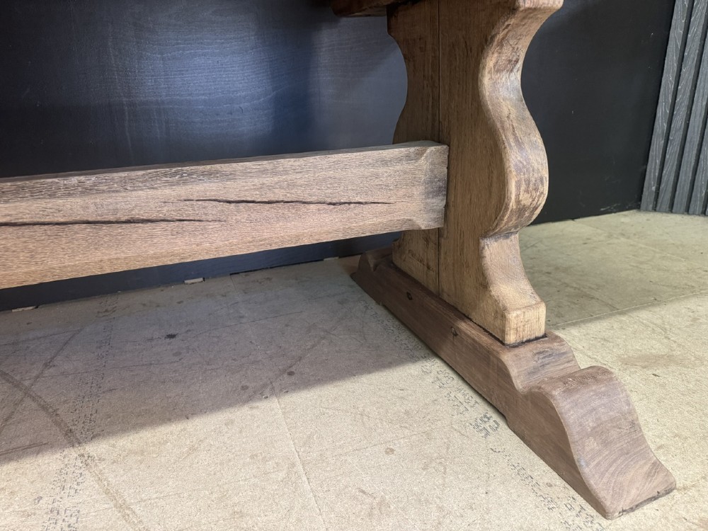 A large oak farmhouse dining table featuring a solid plank top resting on trestle end supports and a keyed stretcher. View XII