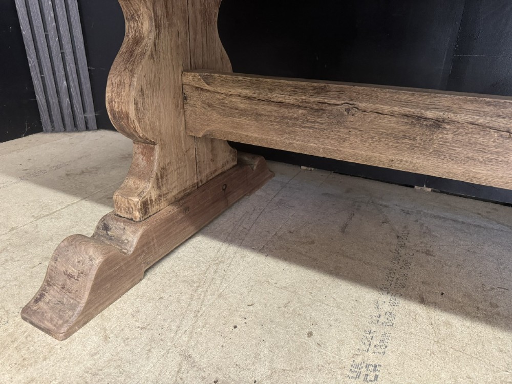 A large oak farmhouse dining table featuring a solid plank top resting on trestle end supports and a keyed stretcher. View XIII