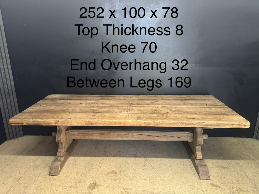 A large oak farmhouse dining table featuring a solid plank top resting on trestle end supports and a keyed stretcher. View XIV