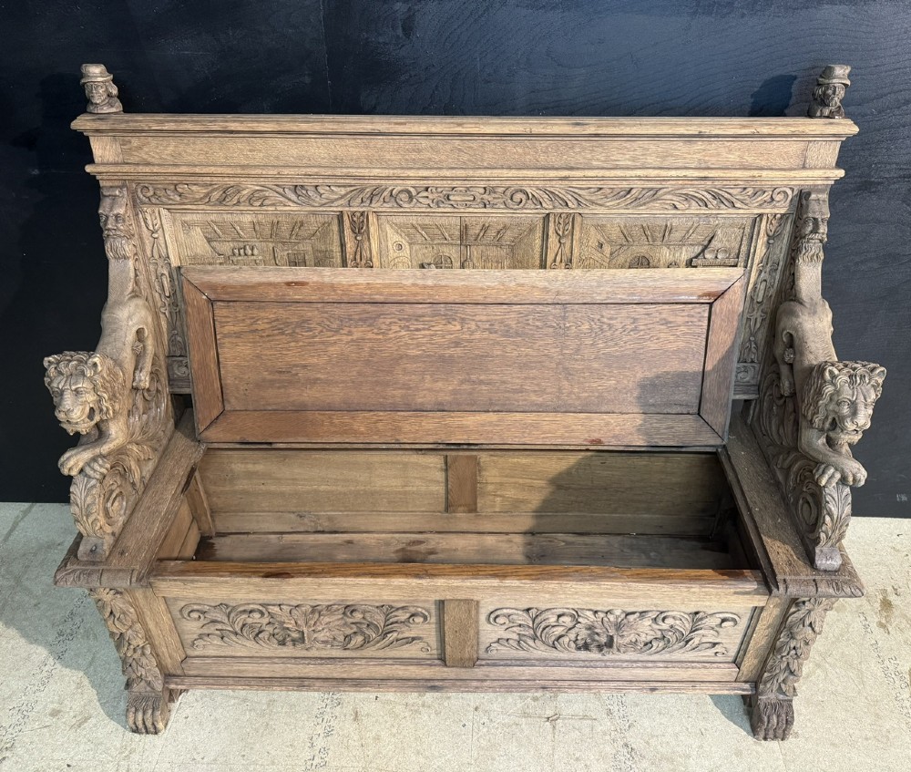 A French carved oak hall bench featuring relief panels depicting tavern scenes, lion figure armrests, and mask carvings on the base. View V