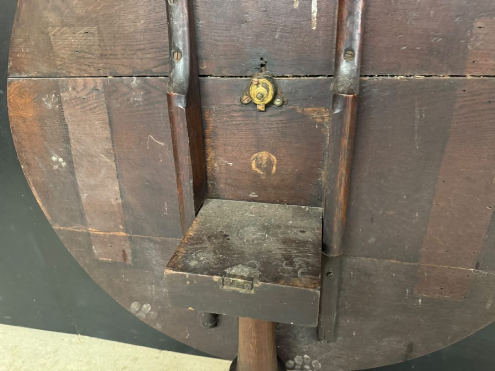 An antique circular oak tilt-top table featuring a three-plank top mounted on a turned pedestal column with three curved tripod legs. View V