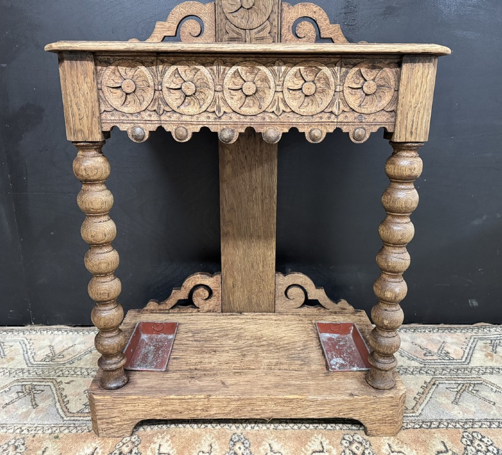 A tall carved oak hall stand featuring two horizontal rails of turned coat pegs, a central column carved with floral rosettes, bobbin-turned legs, and lower umbrella drip trays. View V