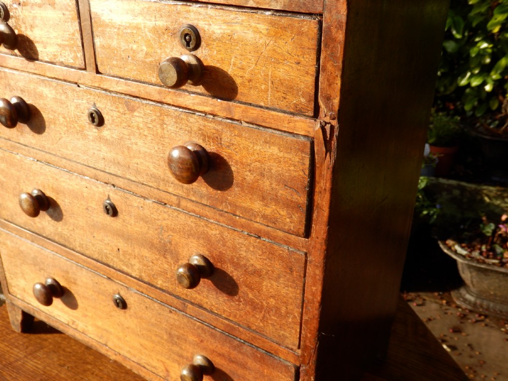A Georgian miniature wooden tabletop chest of drawers with two short over three long drawers, featuring turned timber knobs, brass keyhole escutcheons, and bracket feet. View VI