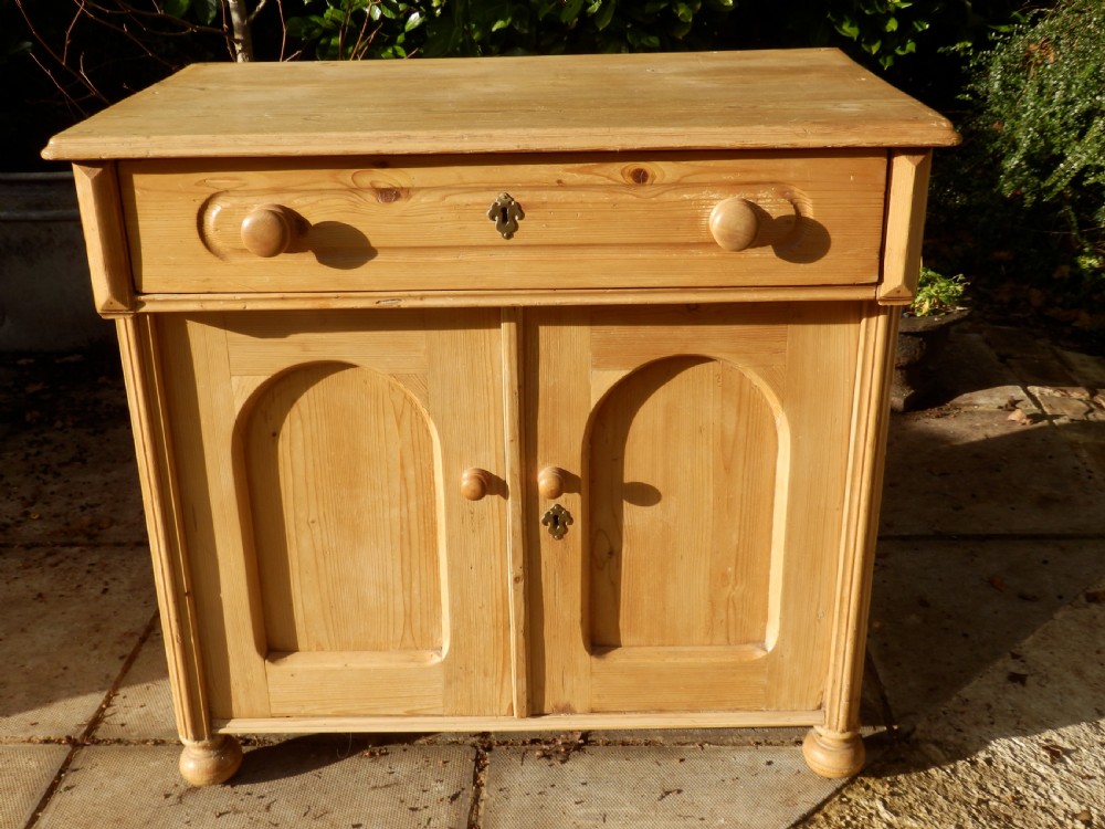 A Victorian pine dresser base with a top drawer, two arched recessed cupboard doors, turned wooden knobs, and brass escutcheons.