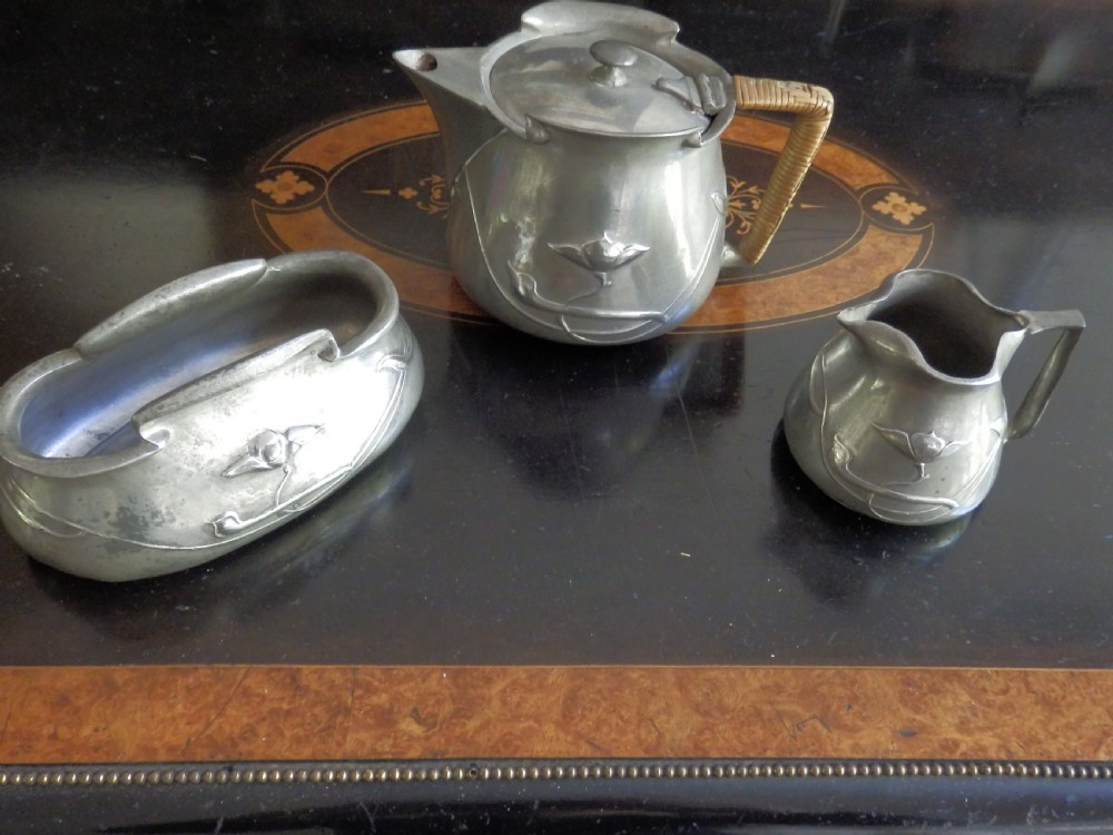 A three-piece pewter tea service consisting of a teapot with a woven cane handle, a sugar bowl, and a cream jug featuring relief plant motifs.