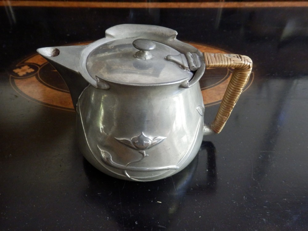 A three-piece pewter tea service consisting of a teapot with a woven cane handle, a sugar bowl, and a cream jug featuring relief plant motifs. View III