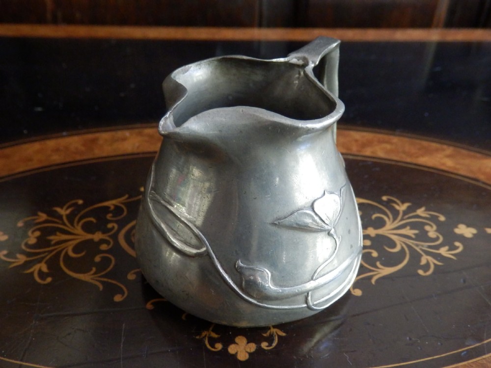 A three-piece pewter tea service consisting of a teapot with a woven cane handle, a sugar bowl, and a cream jug featuring relief plant motifs. View X