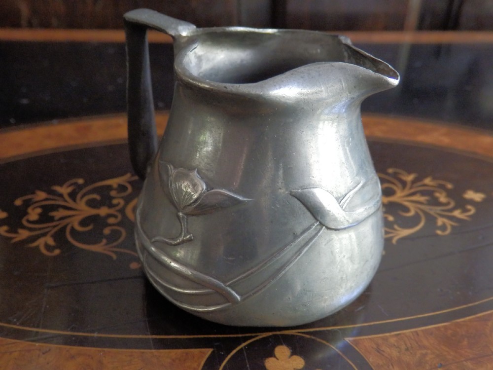 A three-piece pewter tea service consisting of a teapot with a woven cane handle, a sugar bowl, and a cream jug featuring relief plant motifs. View XI